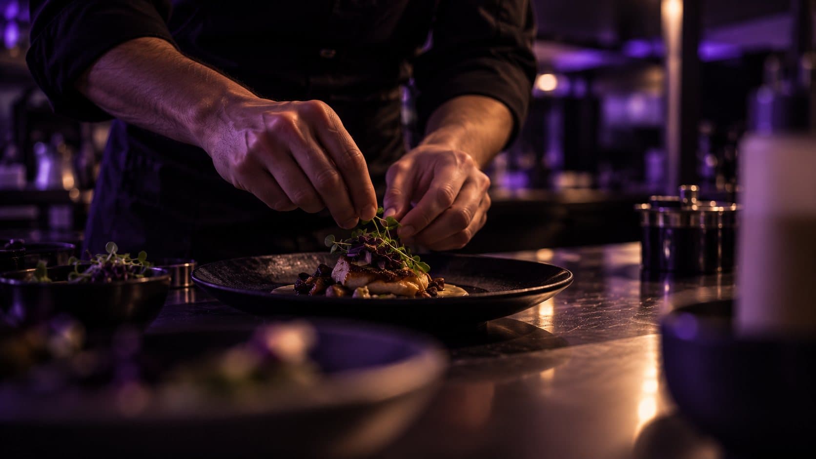 Hände eines Kochs richten ein Gericht auf einem dunklen Teller an, violettes Licht im Hintergrund.